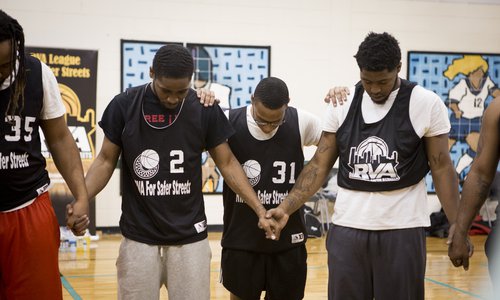 A group of men hold hands with their heads bowed down while on an indoor basketball court